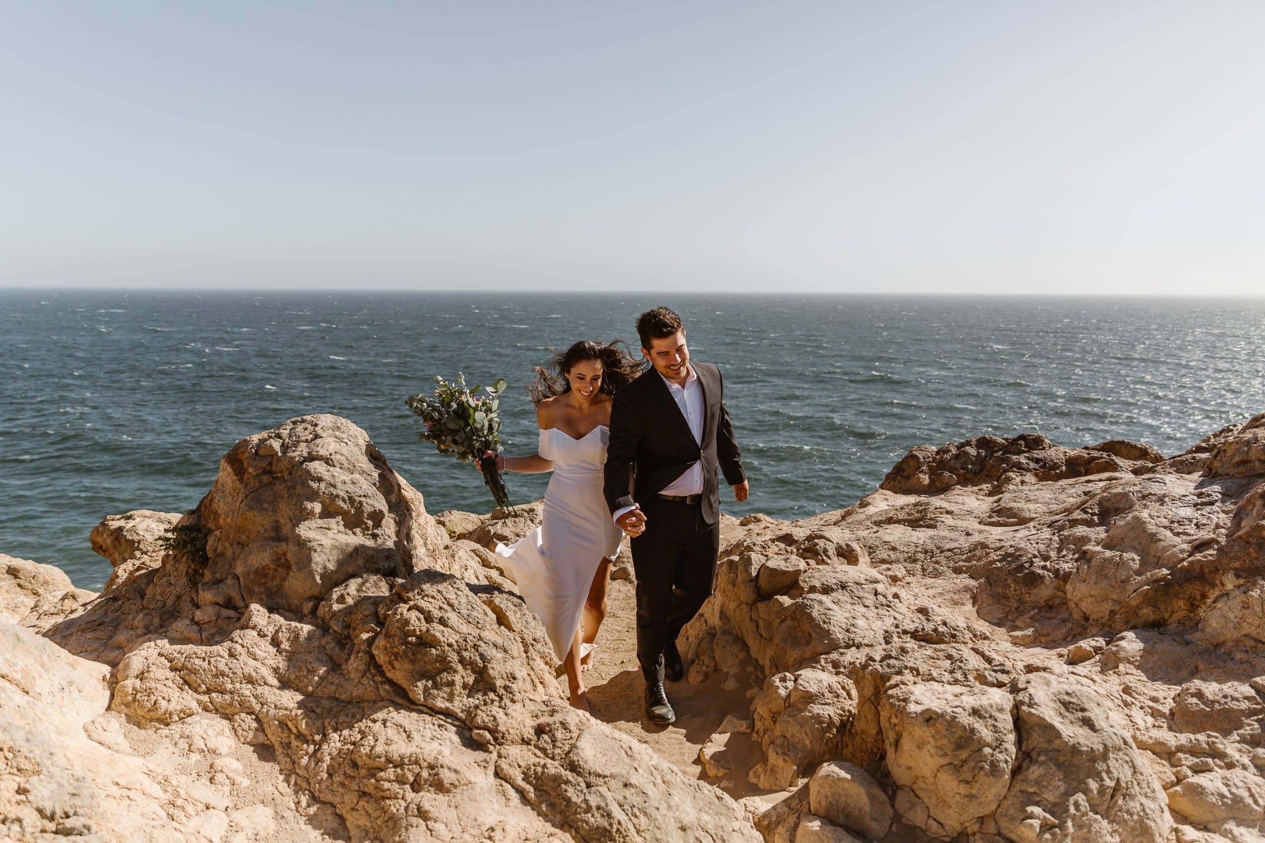 A couple running back from saying their vows, smiling and laughing with the ocean behind them in Malibu CA.